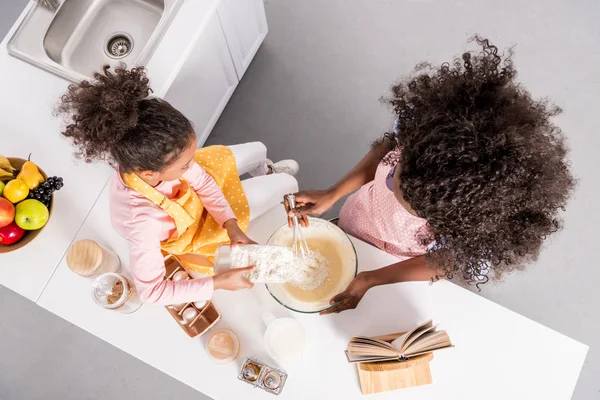 Vista aérea de la madre afroamericana y la hija rizada haciendo masa en un tazón en la cocina - foto de stock