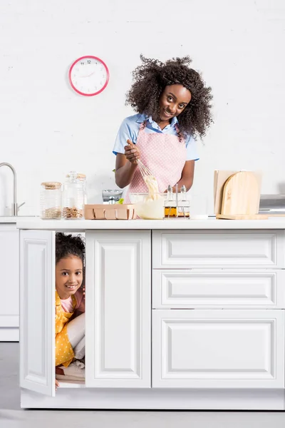 Afroamericana madre haciendo masa mientras hija escondida en cocina - foto de stock