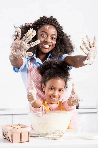 Lächelnd afrikanisch-amerikanische Mutter und lustige Tochter zeigt Hände in Teig und Daumen nach oben auf Küche — Stockfoto