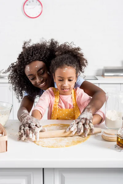 African american mother and daughter rolling dough with pin together on kitchen — Stock Photo