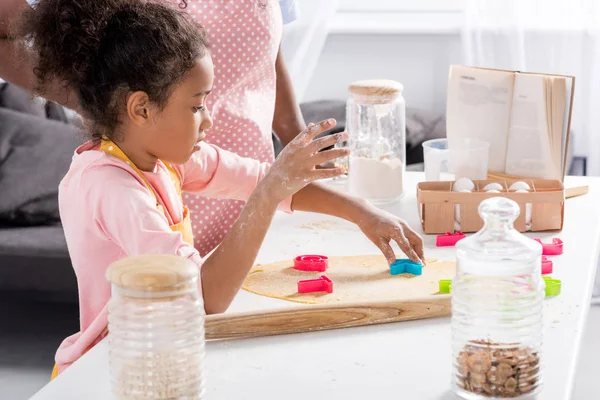 Hija afroamericana haciendo galletas con cortadores de galletas en la cocina - foto de stock