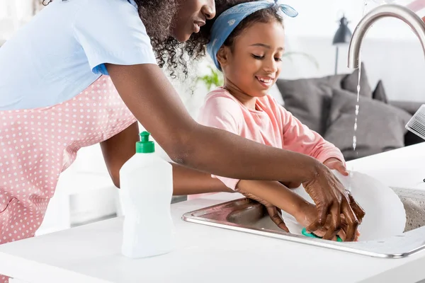 Madre e hija afroamericana lavando platos con detergente en la cocina - foto de stock