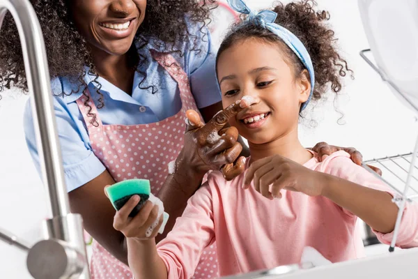 Africano americano madre y hija tener divertido mientras lavado platos con esponja en cocina - foto de stock