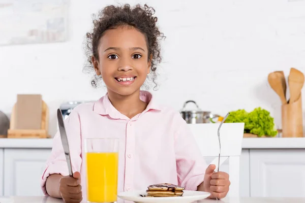 Joyeux gamin afro-américain ayant des crêpes et du jus d'orange pour le petit déjeuner — Photo de stock