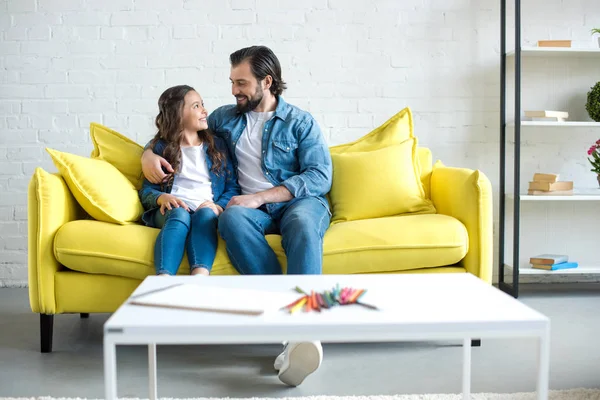 Feliz padre e hija sentados juntos en un sofá amarillo y sonriéndose en casa - foto de stock