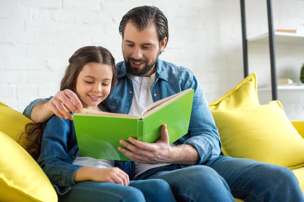 Glücklicher Vater und Tochter lesen zusammen Buch, während sie zu Hause auf dem Sofa sitzen — Stockfoto