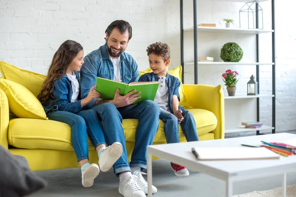 Padre sorridente con i bambini che leggono insieme il libro mentre siedono sul divano di casa — Foto stock