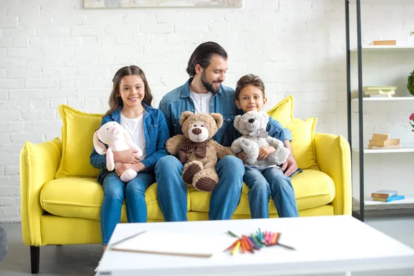 Счастливый отец с очаровательными маленькими детьми, держащими игрушки и сидящими дома на диване — стоковое фото