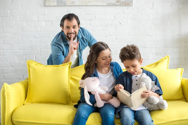 Man with finger on lips smiling at camera while kids reading book on sofa — Stock Photo