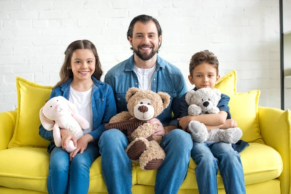 Feliz padre con dos adorables niños sosteniendo juguetes y sonriendo a la cámara en casa - foto de stock