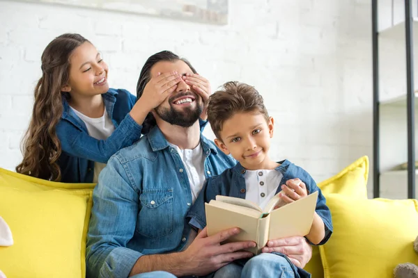 Cute little daughter closing eyes to happy father while father and son reading book on sofa at home — Stock Photo