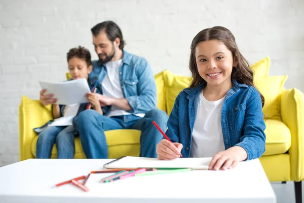 Adorabile disegno bambino con matite colorate e sorridente alla macchina fotografica mentre padre e fratello seduti sul divano dietro — Foto stock