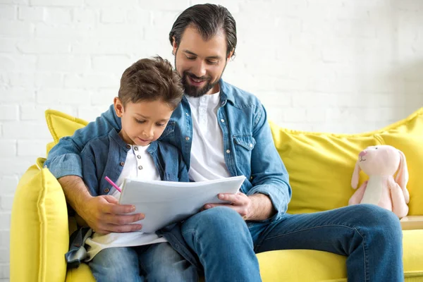 Счастливый отец и милый маленький сын рисует сидя вместе на диване дома — стоковое фото