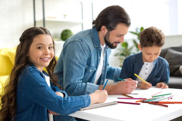Adorable child smiling at camera while drawing with father and brother at home — Stock Photo