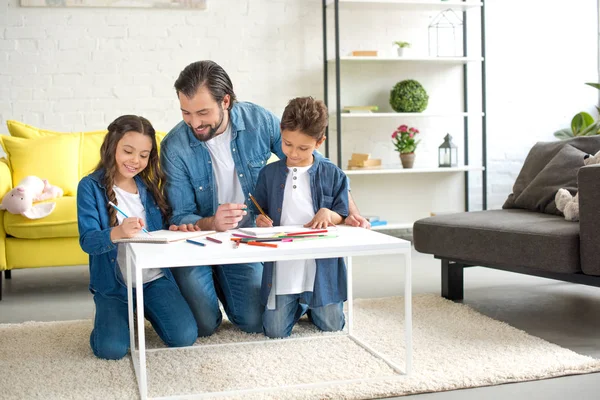 Padre feliz con dos niños arrodillados en la alfombra y dibujo con lápices de colores en casa - foto de stock