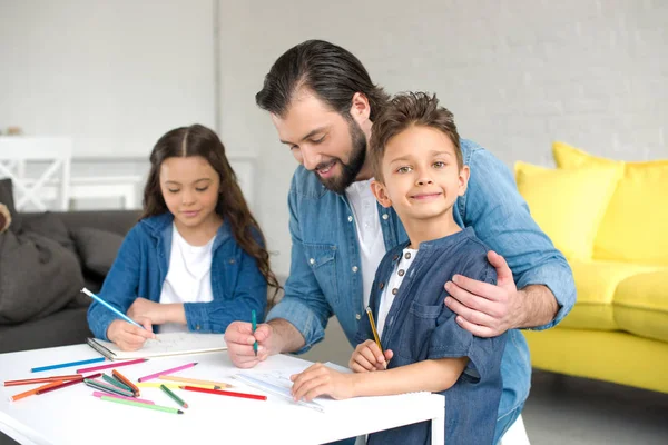 Счастливый отец и двое очаровательных детей рисуют цветными карандашами дома — стоковое фото