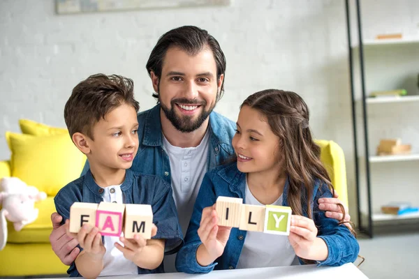 Feliz padre con dos adorables niños sosteniendo cubos con la palabra familia - foto de stock