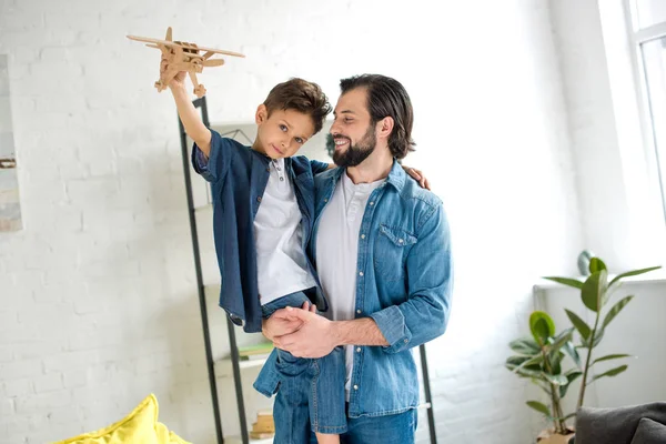 Feliz padre llevando lindo pequeño hijo jugando con juguete avión y mirando a la cámara - foto de stock