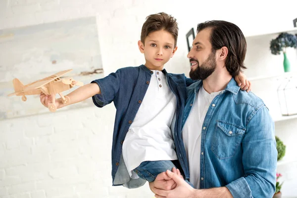 Feliz padre llevando adorable pequeño hijo jugando con juguete avión y mirando a la cámara en casa - foto de stock