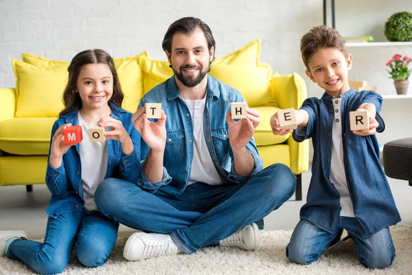 Happy father and two kids holding cubes with word mother and smiling at camera — Stock Photo
