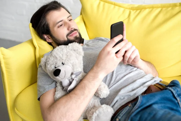 Усміхнений молодий чоловік використовує смартфон, лежачи на дивані і обіймаючи плюшевого ведмедя — стокове фото