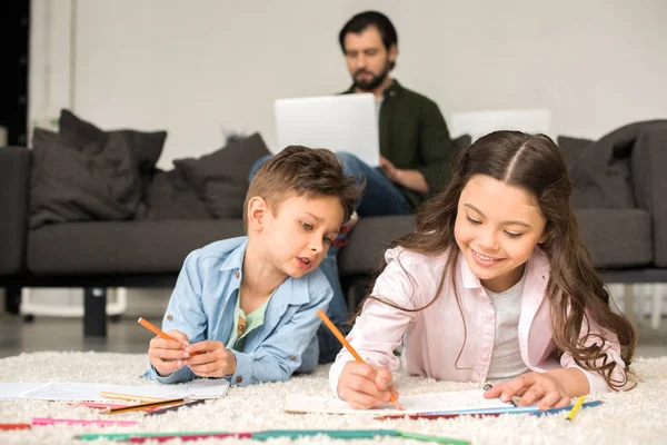 Enfants souriants mignons couchés sur le tapis et le dessin avec des crayons de couleur tandis que le père en utilisant un ordinateur portable derrière — Stock Photo