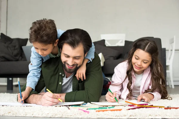 Happy father with cute children lying on carpet and drawing with colored pencils — Stock Photo