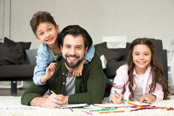 Glücklicher Vater mit niedlichen kleinen Kindern, die in die Kamera lächeln, während sie zu Hause mit Buntstiften zeichnen — Stockfoto