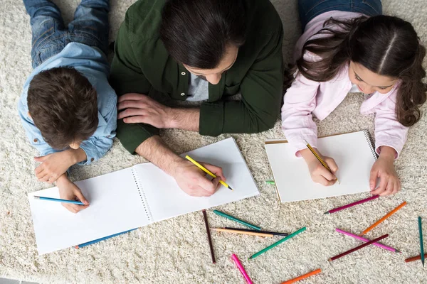 Вид сверху на отца с детьми, рисующими цветными карандашами дома — стоковое фото