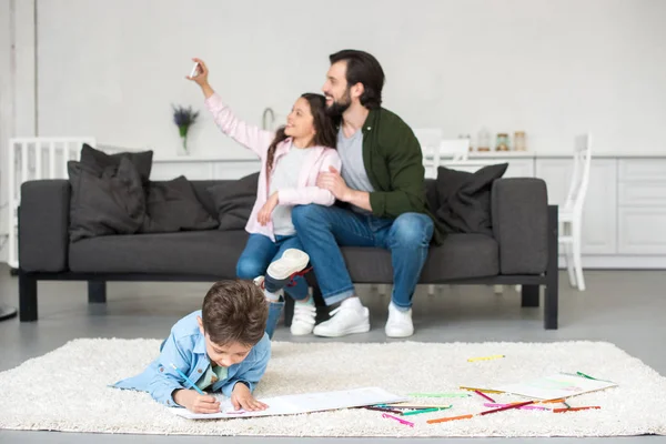 Happy father and daughter sitting on sofa and taking selfie with smartphone while little boy drawing on carpet at home — Stock Photo