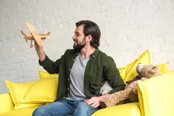 Усміхнений молодий чоловік сидить на дивані і грає з дерев'яним іграшковим літаком — стокове фото
