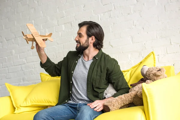 Smiling young man sitting on sofa and playing with wooden toy plane — Stock Photo