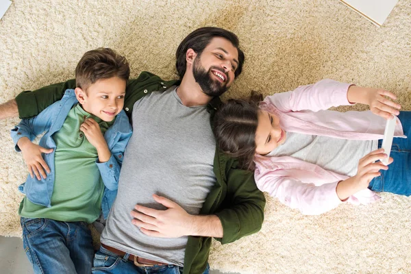 Top view of father with two adorable kids lying on carpet and taking selfie with smartphone — Stock Photo