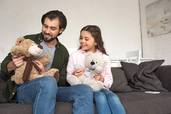 Feliz pai e filha sentados no sofá e brincando com ursinhos de pelúcia em casa — Fotografia de Stock