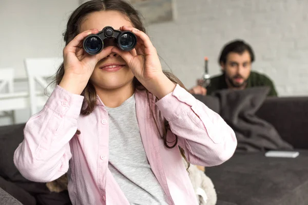 Mignon sourire enfant jouer avec jumelles tandis que père avec jouet pistolet caché derrière canapé — Photo de stock