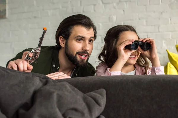 Улыбающиеся отец и дочь с биноклем и игрушечным пистолетом прячутся за диваном и играют вместе дома — стоковое фото