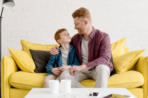 Feliz pelirroja padre e hijo sentado juntos en el sofá y sonriendo el uno al otro en casa - foto de stock