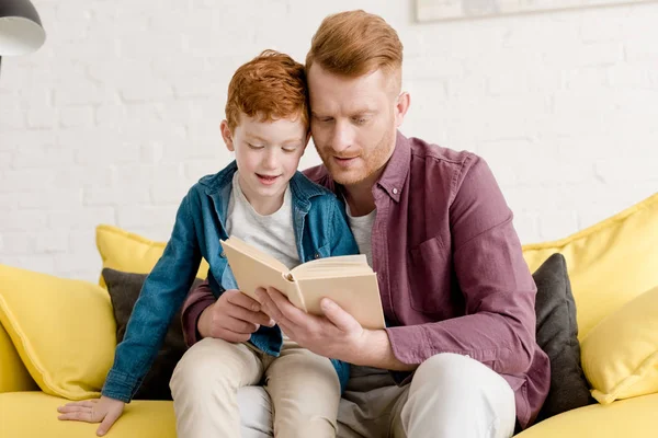 Pai e filho sentados no sofá e lendo livro juntos — Fotografia de Stock