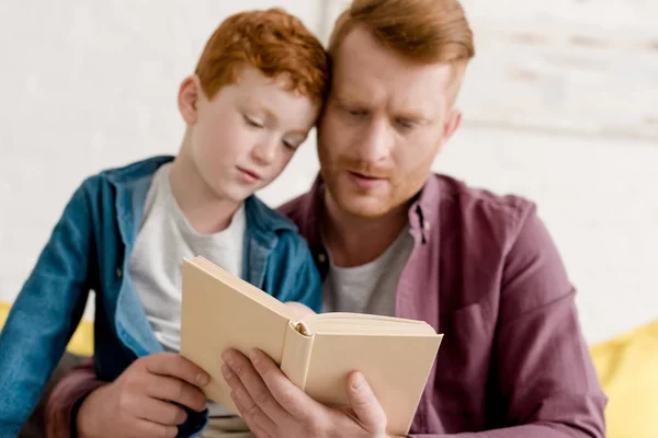 Vista de primer plano del padre y el hijo enfocados leyendo el libro juntos en casa - foto de stock