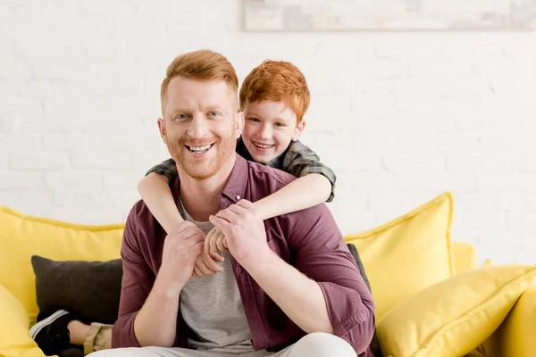 Feliz pelirroja padre e hijo abrazando y sonriendo a la cámara en casa - foto de stock