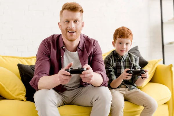 Happy father and son sitting on sofa and playing with joysticks at home — Stock Photo