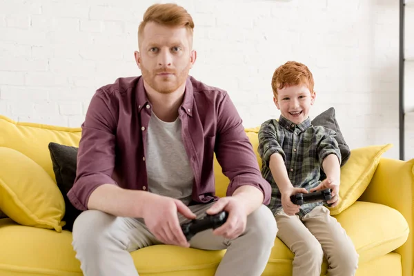 Padre e hijo sentados en el sofá y jugando con joysticks en casa - foto de stock