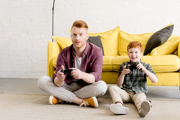 Feliz padre e hijo sentado en la alfombra y jugando con joysticks en casa - foto de stock