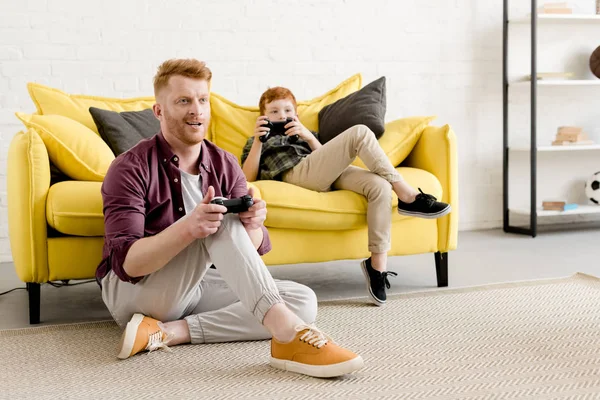 Sorrindo de cabelos vermelhos pai e filho brincando com joysticks juntos em casa — Fotografia de Stock