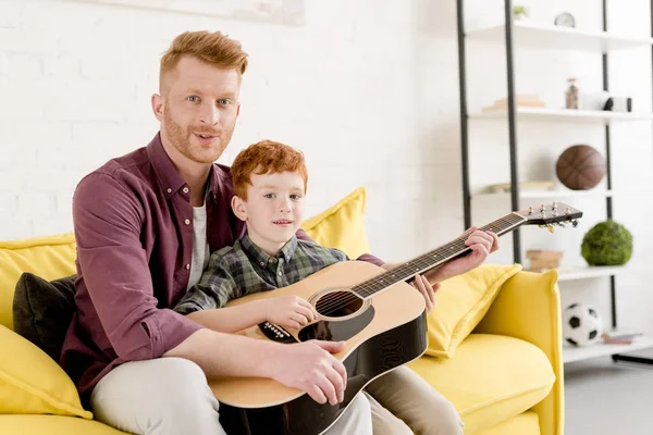 Heureux père et fils tenant guitare acoustique et souriant à la caméra à la maison — Photo de stock