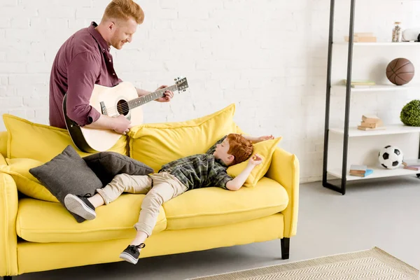 Padre sonriente tocando la guitarra y lindo hijo pequeño acostado en el sofá - foto de stock