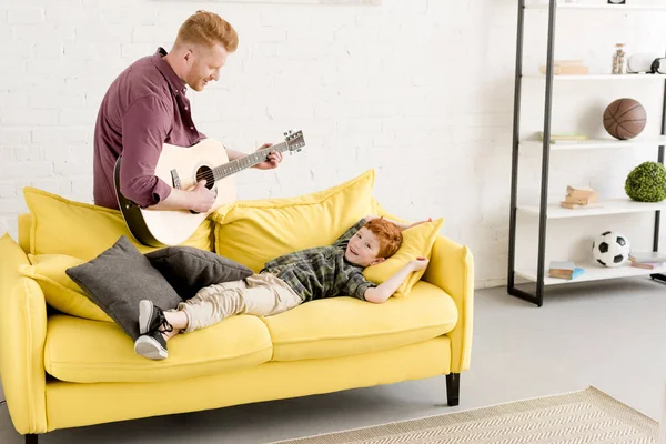 Счастливый отец играет на гитаре и милый улыбающийся маленький сын лежит на диване — стоковое фото