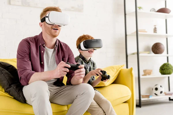 Happy father and son in virtual reality headsets playing with joysticks at home — Stock Photo