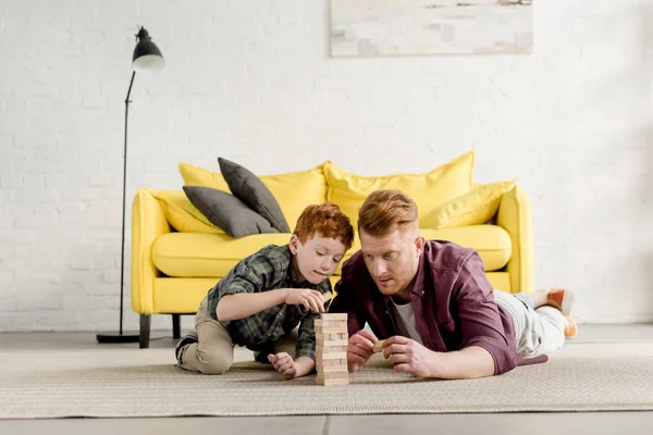 Concentré rousse père et fils couché sur le tapis et jouer avec des blocs de bois à la maison — Photo de stock