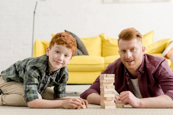 Heureux père et fils roux souriant à la caméra tout en jouant avec des blocs de bois à la maison — Photo de stock